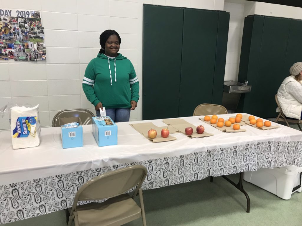 A Girl Scout standing by her healthy table offering apples and oranges
