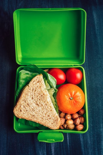 A green lunchbox with a healthy sandwich, orange, nuts, and a couple of small tomatoes